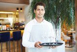 Portrait of young waiter in uniform holding tray with empty wineglasses standing at restaurant