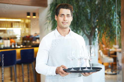 Portrait of young waiter in uniform holding tray with empty wineglasses standing at restaurant