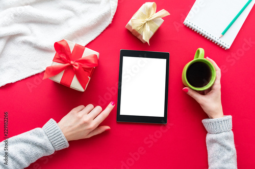 Female hands, woman holding a Cup of coffee or tea, girl uses the tablet pc. Red table, gifts boxes for the holidays, background with copy space for advertisement, top view