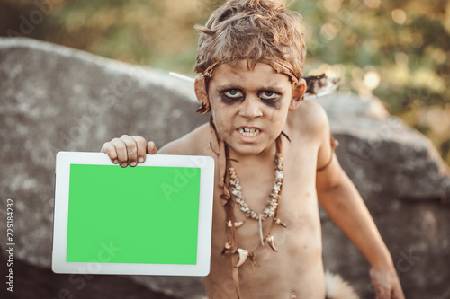 Caveman, manly boy holding tablet PC with green screen. Funny young primitive boy outdoors. Evolution degradation concept. Calm boy outside against rocky background. Prehistoric tribal man outside on