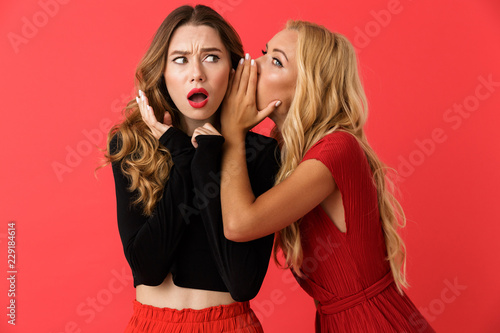 Shocked young friends women standing isolated over red background gossiping.