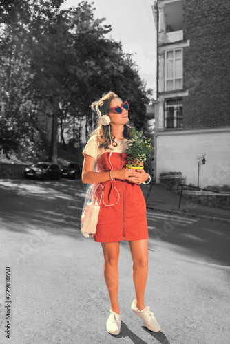 Going with plant. Slim and beautiful woman wearing red dress and white sneakers listening to music while going home with plant
