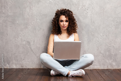 Foto Happy young woman sitting on the floor with crossed legs and using laptop on gray background