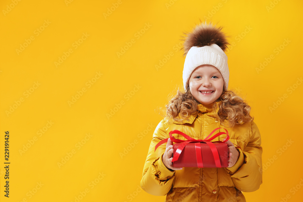 Happy little girl preschooler in a winter jacket and  in a knitted hat is holding a box with a gift. The concept of giving and receiving gifts. Yellow on a yellow background, red.