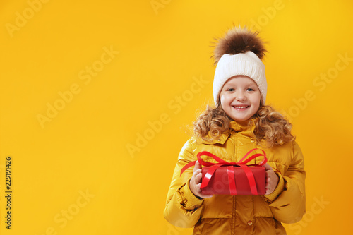 Happy little girl preschooler in a winter jacket and in a knitted hat is holding a box with a gift. The concept of giving and receiving gifts. Yellow on a yellow background, red.