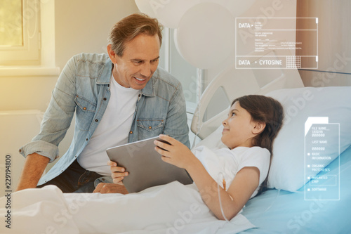 Modern device. Emotional ill girl looking curiously at her cheerful father while staying in a hospital bed with a modern tablet in her hands