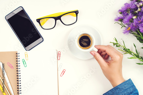 Female hand with coffee cup. Top view of work desk. Coffee, glasses, notebook, pencil, glasses, paper clips and mobile phone on white table. Mockup. Freelance feminine workspace. Morning time at work.