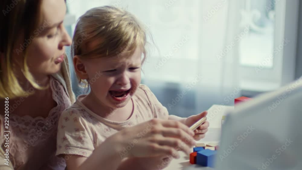 of young mother holding crying toddler girl on her laps and showing something on mobile phone while trying to calm her down of young mother holding crying toddler girl on her laps and showing something on mobile phone while trying to calm her down