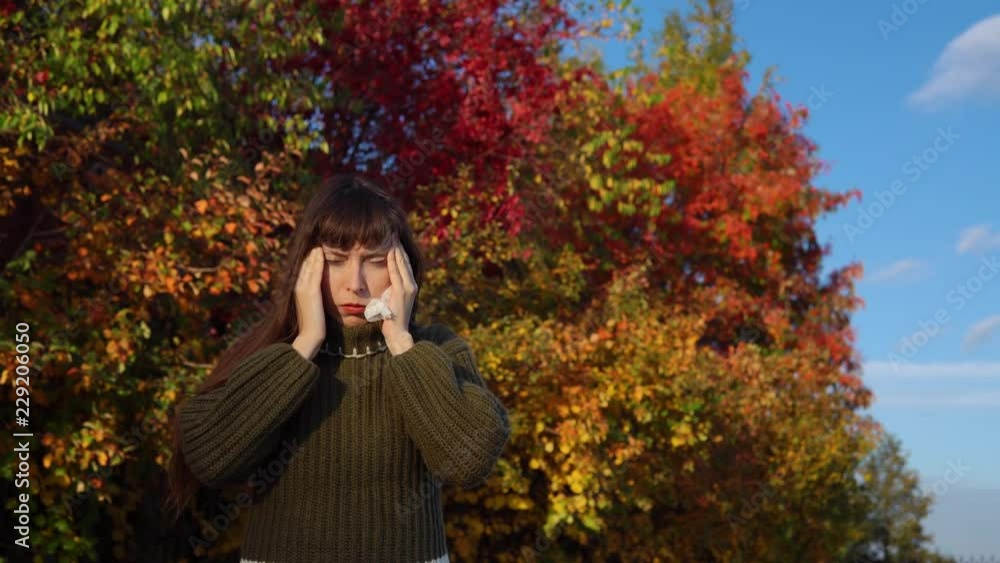 A young sick woman in a green knitted sweater strokes her head against the background of multi-colored foliage in the city park in Indian summer. A young sick woman in a green knitted sweater strokes her head against the background of multi-colored foliage in the city park in Indian summer.