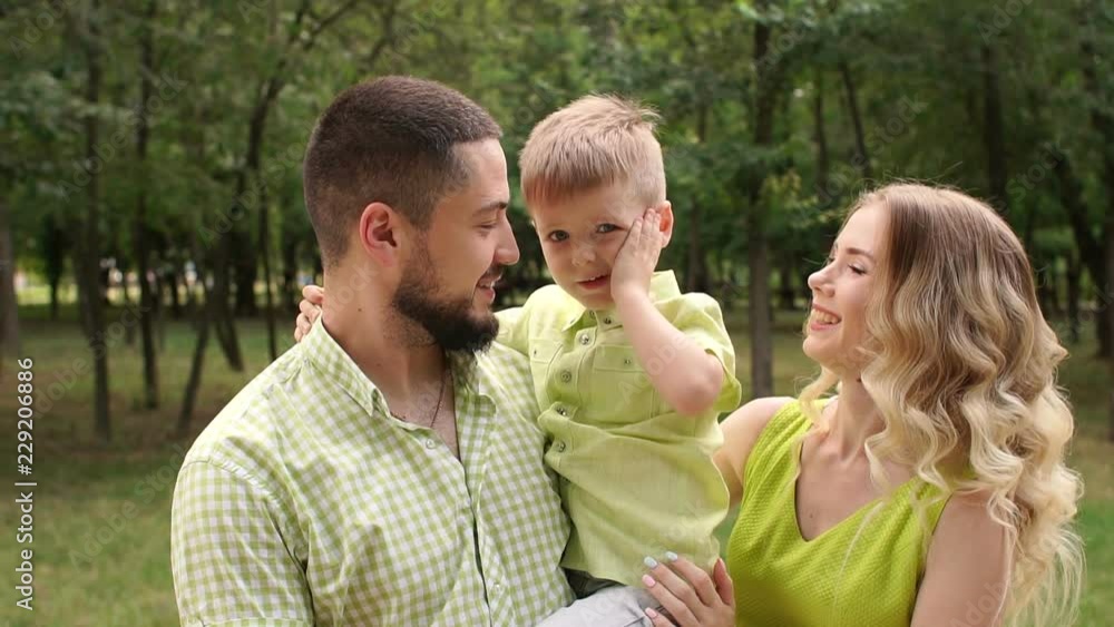 Close-up of a happy smiling little boy in the hands of a loving mom and dad in the park. Loving parents kiss their son outdoors in the summer park. Close-up of a happy smiling little boy in the hands of a loving mom and dad in the park. Loving parents kiss their son outdoors in the summer park.
