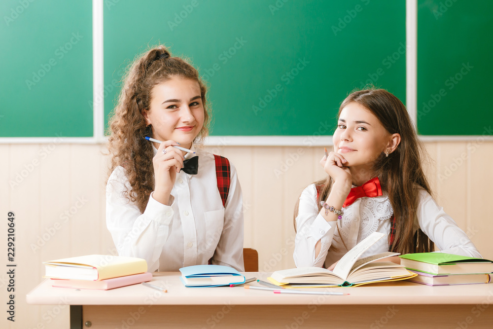 2 schoolgirls in school uniform sit on the background of a green board at the desk with books 2 schoolgirls in school uniform sit on the background of a green board at the desk with books