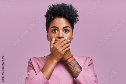 Terrified dark skinned cute young female with Afro hairstyle, covers mouth with great fear, tries not to cry, has scared shocked expression, wears striped sweater, isolated lavender background photo