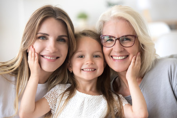 Portrait of three generations of women look at camera posing for family picture, cute little girl hug mom and granny enjoy time at home, smiling mother, daughter and grandmother spend weekend together