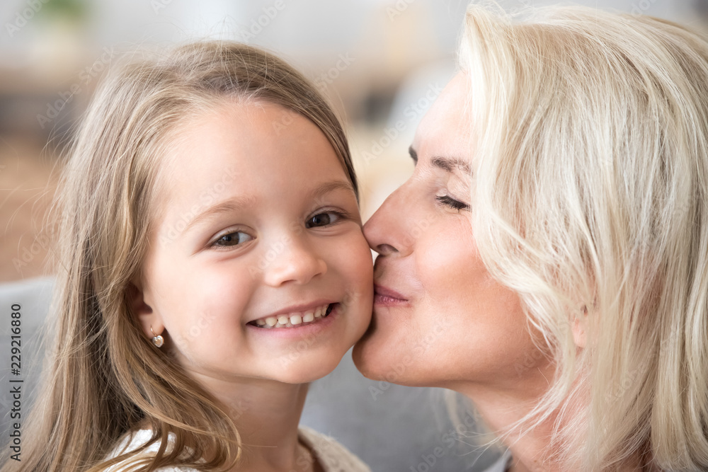 Portrait of loving grandmother kissing granddaughter on cheek showing care and affection, small cute grandchild look at camera spending time with granny at home cuddling and hugging. Family concept Portrait of loving grandmother kissing granddaughter on cheek showing care and affection, small cute grandchild look at camera spending time with granny at home cuddling and hugging. Family concept