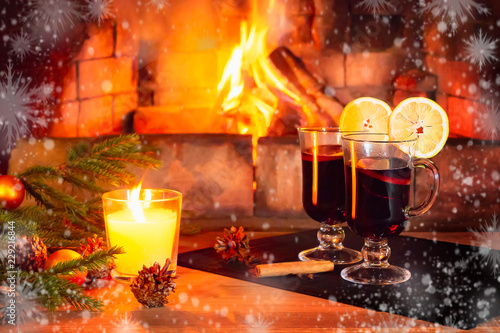 Two glasses with mulled wine, a candle, fir branches with decorations on a wooden table against the background of a burning fireplace framed by hoarfrost and snowflakes. Romantic christmas concept