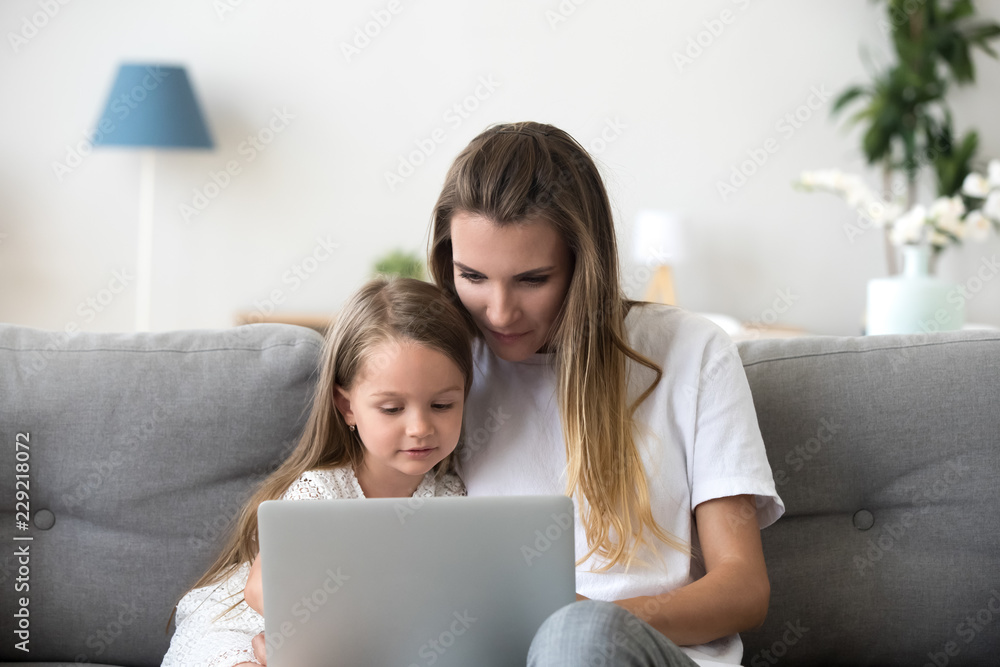 Mother and daughter sit on couch using laptop watching video or cartoon online, cute little girl spend time with mom, relax at home browsing computer or shopping together. Young and technology concept Mother and daughter sit on couch using laptop watching video or cartoon online, cute little girl spend time with mom, relax at home browsing computer or shopping together. Young and technology concept