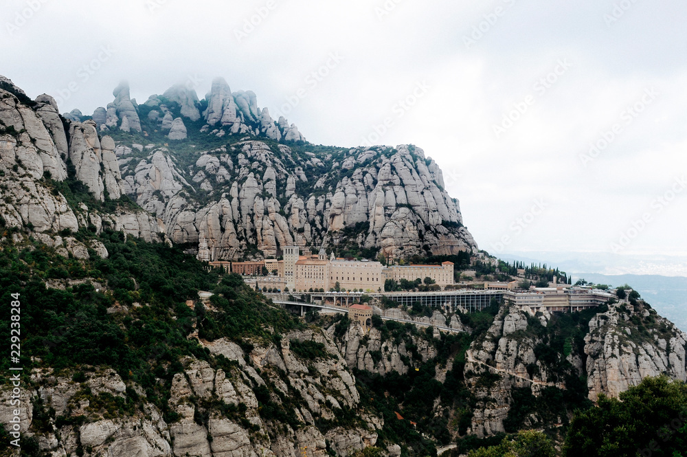 Montserrat, Catalonia, Spain. Top View Of Hillside Cave Santa Cova De Montserrat Or Holy Cave Of Montserrat In Summer Day. Santa Maria De Montserrat Abbey.
