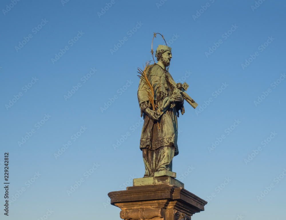 Baroque Statue of Saint John of Nepomuk with Golden Star Halo holding crucifix with Jesus Christ on Charles Bridge in Prague, Czech Republic, sunny day, clear blue sky