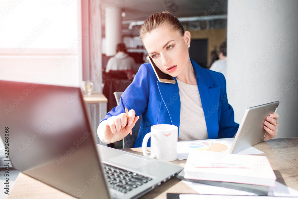 A picture of serious woman working in office. She is holding a tablet in one hand and a pen in the other one. Girl is looking at the laptop's screen. Also she has a cup of tea in fromnt of her. A picture of serious woman working in office. She is holding a tablet in one hand and a pen in the other one. Girl is looking at the laptop's screen. Also she has a cup of tea in fromnt of her.