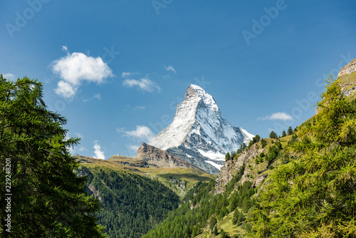 Scenic summer view on snowy Matterhorn peak in sunny day with green meadows, pine tree forests, hills, blue sky and white clouds in background in Switzerland photo