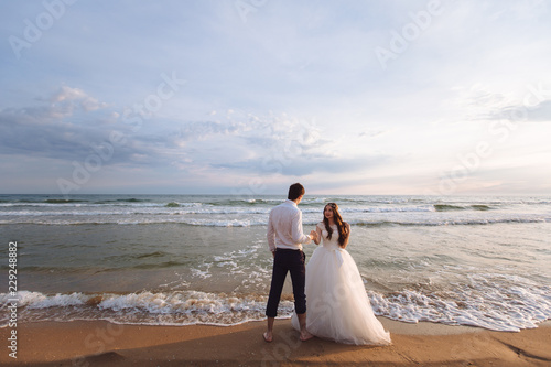A beautiful couple of newlyweds  the bride and groom walking on the beach. Gorgeous sunset and sky. Wedding dresses  a white luxury dress for a girl. Family concept  honeymoon.