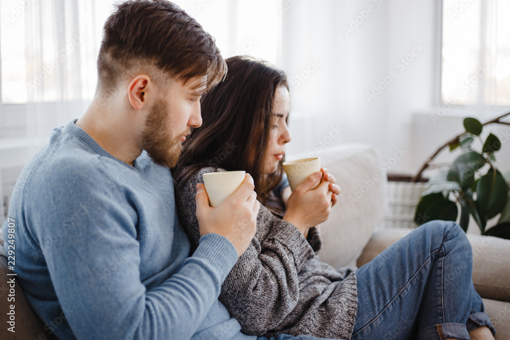 Young couple relaxing at home sitting at sofa with hot drinks. Warming, calmness, home owning, comfort concept Young couple relaxing at home sitting at sofa with hot drinks. Warming, calmness, home owning, comfort concept