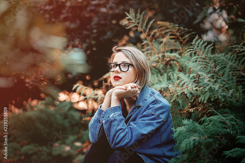 beautiful young girl in jacket and glasses sitting on a gorund in a park. Fall season time scene. Hipster trends clothes style photo