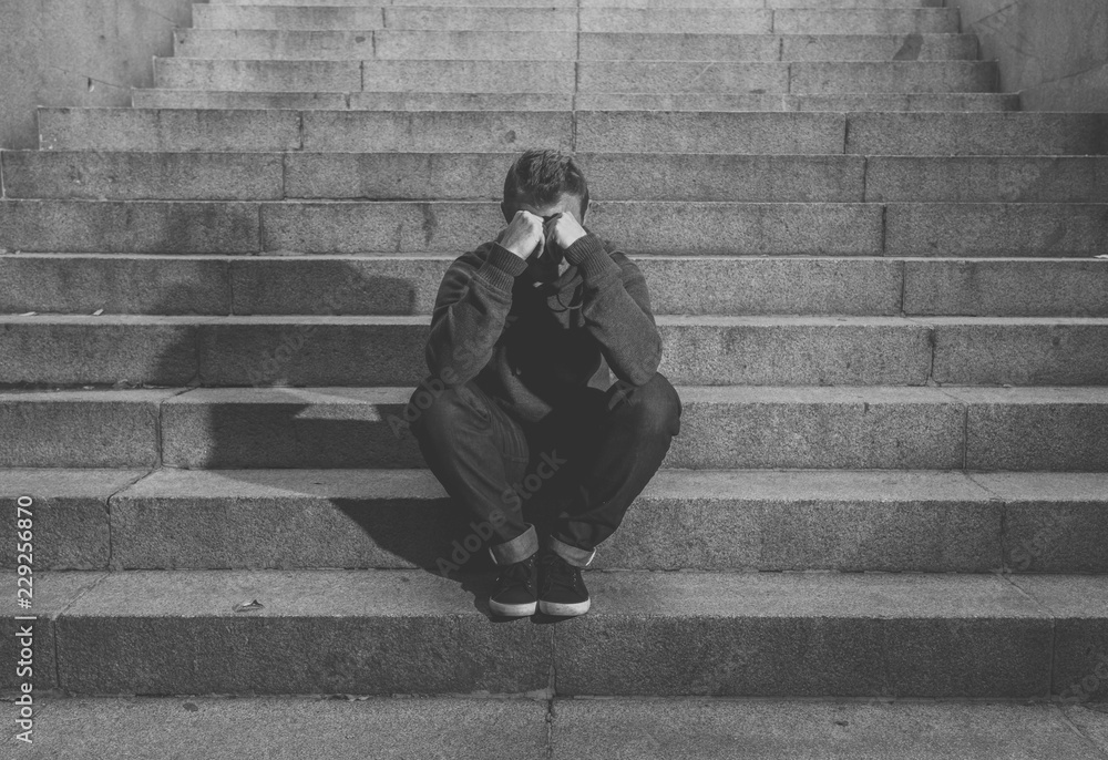 Depressed sad young man crying sitting on stairs feeling miserable lonely in urban scene