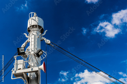 Close-up of a mast with stay cables, lamps and a horn on a cruise ship against blue sky and white clouds, small white metal ladder, sunny day