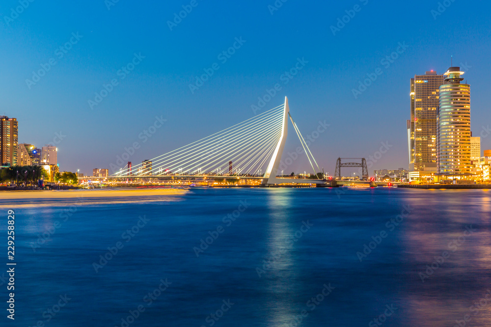 Obraz premium Cityscape on a calm night with illuminated Erasmus cable-stayed bridge over the river Maas by buildings against clear blue sky, yellow lights, Rotterdam in the Netherlands. Long exposure photography