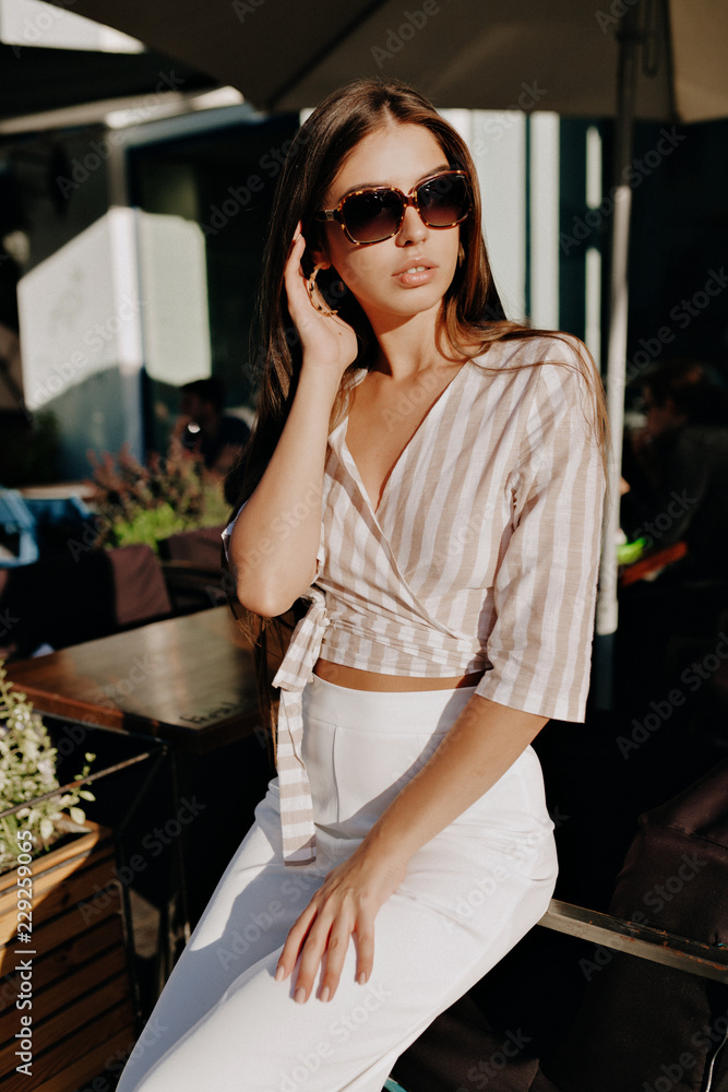 Stylish modern model in sunglasses and fashion t-shirt and white trouthers posinf in sunlight outdoor in warm weather. Photo of fashionable girl with beautiful brown hair smiling to camera.