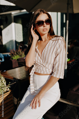 Obraz na płótnie Stylish modern model in sunglasses and fashion t-shirt and white trouthers posinf in sunlight outdoor in warm weather. Photo of fashionable girl with beautiful brown hair smiling to camera. w salonie