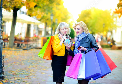 Two happy girls in sunglasses with shopping bags, use a smartphone and smile. positive emotions