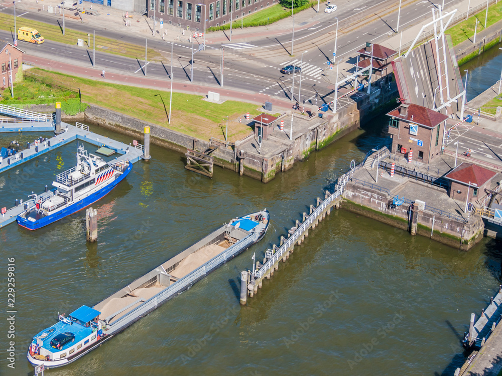 Aerial view of a cargo ship sailing on New Maas river towards a canal with raised drawbridge, boat anchored at pier, vehicular street in background, sunny day in Rotterdam, South Holland, Netherlands