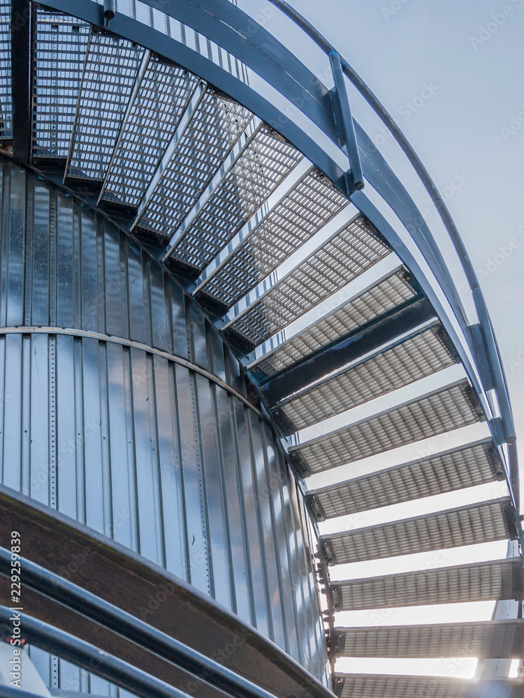 Fototapeta premium Low angle perspective of exterior metal spiral or elliptical stairs with railing against a sheet metal wall in a tower, grating tread or galvanized steel grille, sunny day with a blue sky