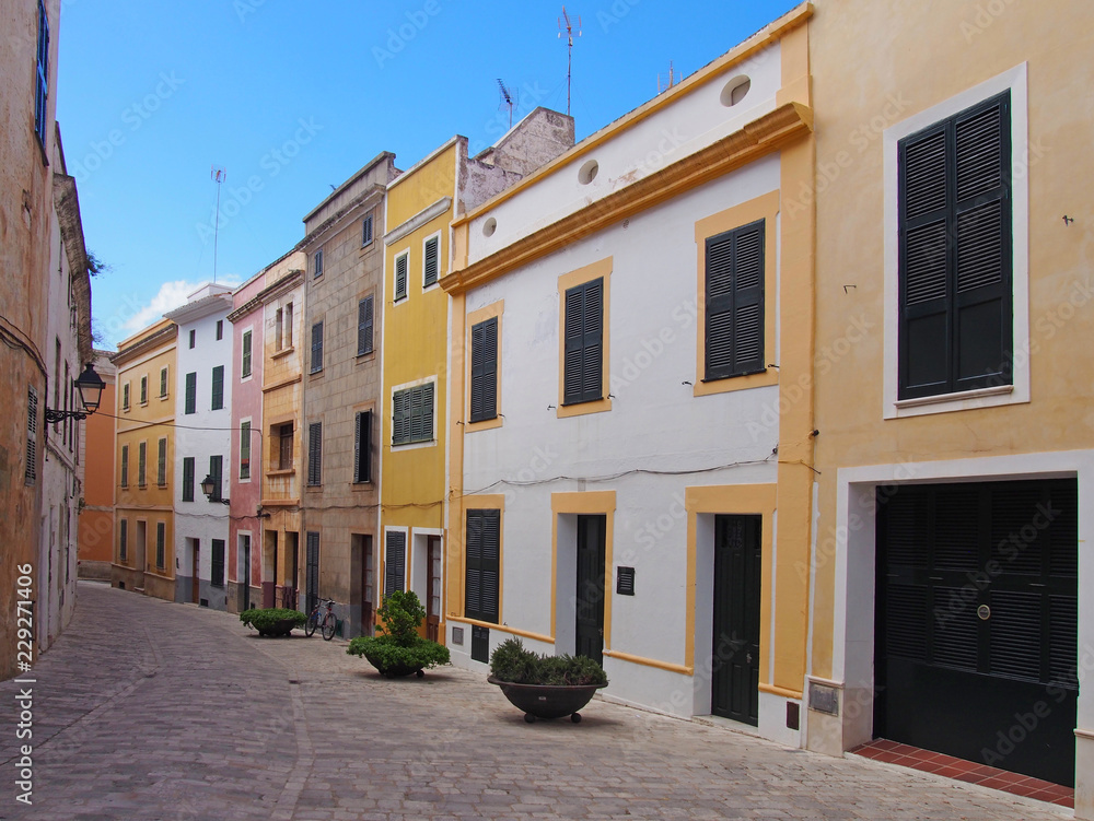a scenic view of an empty street of bright painted yellow traditional houses in ciutadella menorca with bright blue summer sky