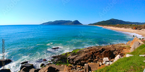 Caldeirão and Armação beaches in Florianopolis, Brazil, during a sunny morning. Rocks can be seen close to the photographer, and mountains can be seen far away. photo