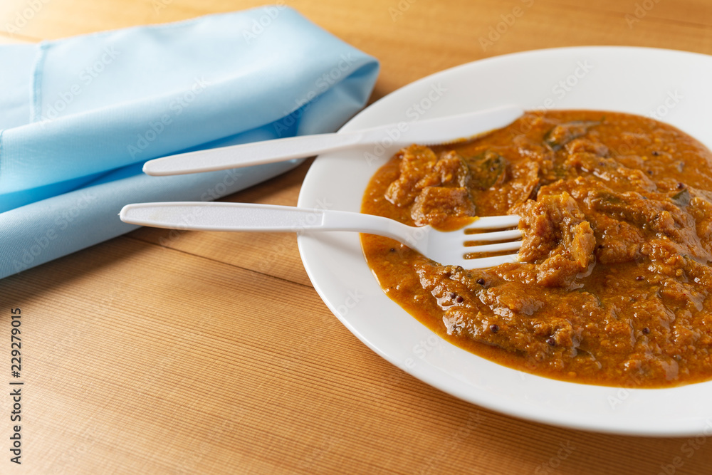 A meal of eggplant curry on a white plate with a plastic fork and knife in the food plus a blue napkin to the side atop a wood table top. A meal of eggplant curry on a white plate with a plastic fork and knife in the food plus a blue napkin to the side atop a wood table top.