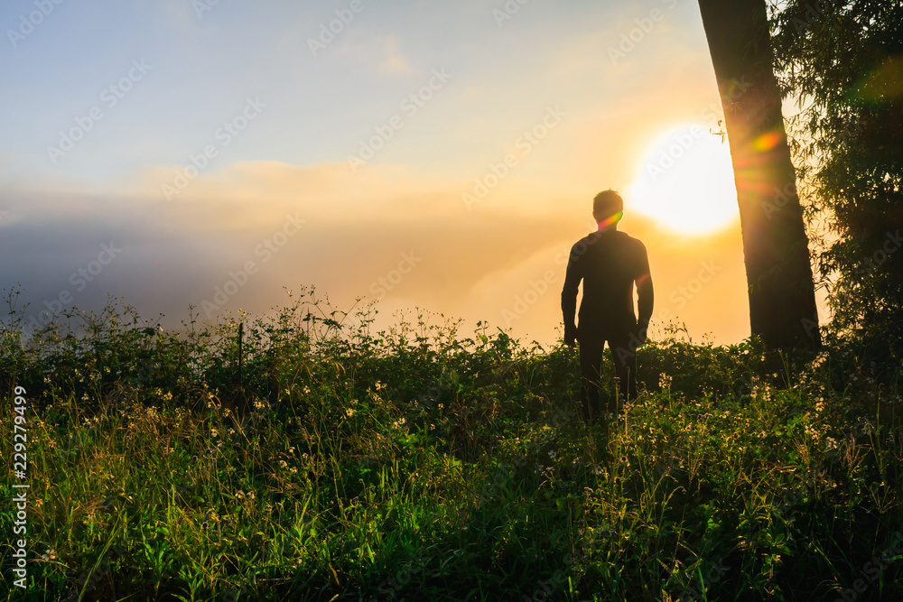 Silhouette of man standing on top mountain and looks into the distance at morning sunrise with foggy mountain landscape