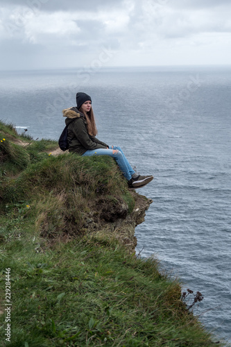 Girl sits on the edge of the steep Cliffs of Moher in Ireland