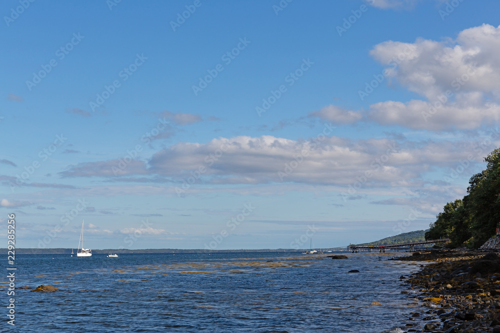 The rocky shoreline of Belfast, Maine on a cloudy summer day with distant boats at their moorings and clouds above.
