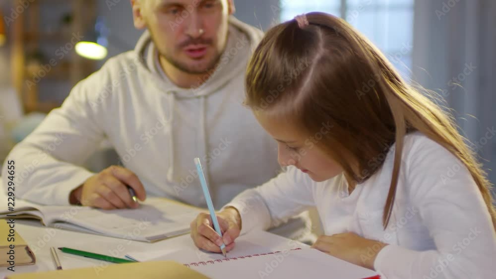 Man helping daughter with study at home: cute girl writing down with pencil while father reading textbook and telling how to do homework Man helping daughter with study at home: cute girl writing down with pencil while father reading textbook and telling how to do homework