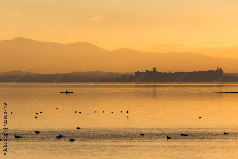 Beautiful view of Trasimeno lake (Umbria, Italy) at sunset, with orange tones, birds on water, a man on a canoe and Castiglione del Lago town on the background