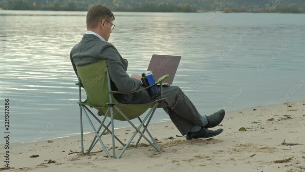 Mature businessman sitting and relaxing near a river. Man in suit and using laptop.