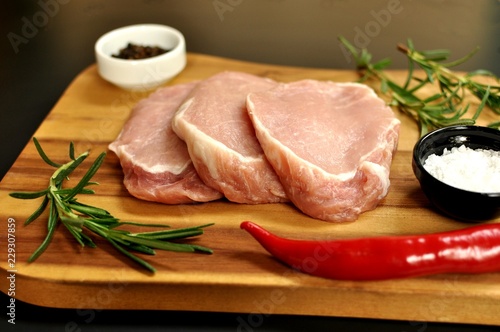 Raw fresh uncooked sliced pork fillet dish with rosemary, pepper, salt, red chili pepper, on wooden board and black background. Top side view