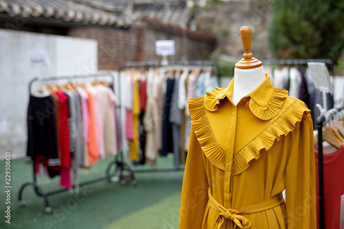 Tableau sur toile Yellow second hand dress is put on mannequin in front of the house together with many used clothes in the background