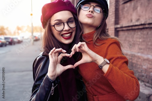 Outdoors fashion portrait young pretty best girls friends in friendly hug. Walking at the city. Posing at the street. Wearing stylish outerwear and hats. Bright make up. Positive emotions. photo