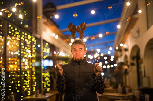 People, holidays and christmas concept - surprised man in deer's horns holding two bengals light or sparklers outdoors © satura_
