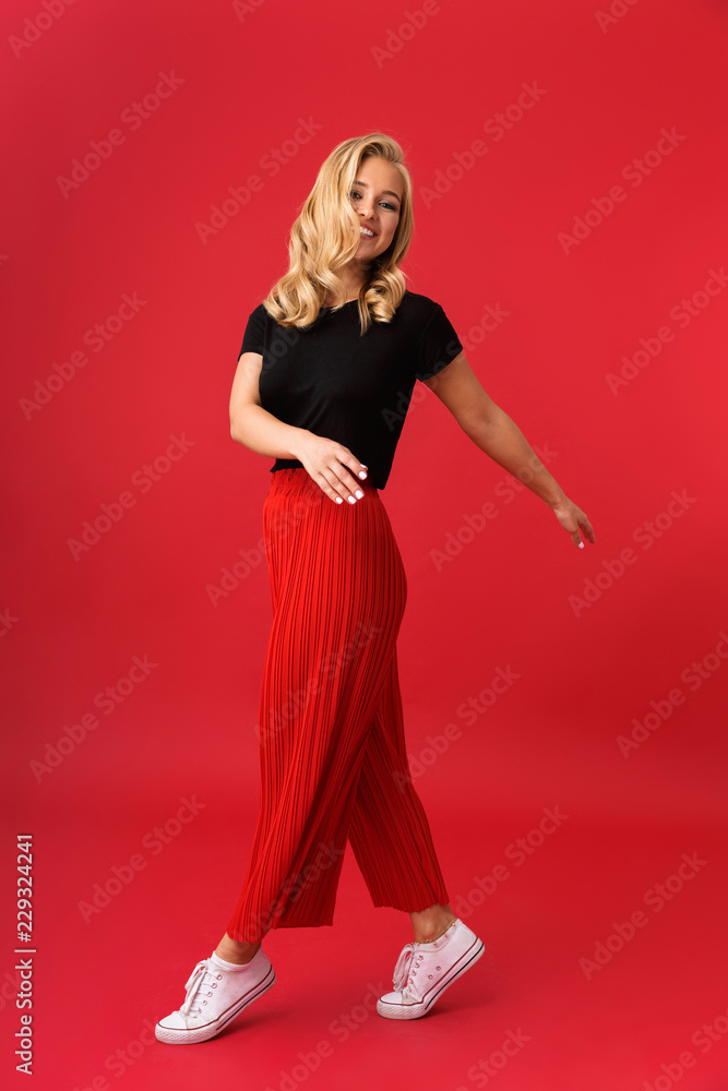 Full length portrait of caucasian blond woman 20s smiling and walking, isolated over red background in studio Full length portrait of caucasian blond woman 20s smiling and walking, isolated over red background in studio