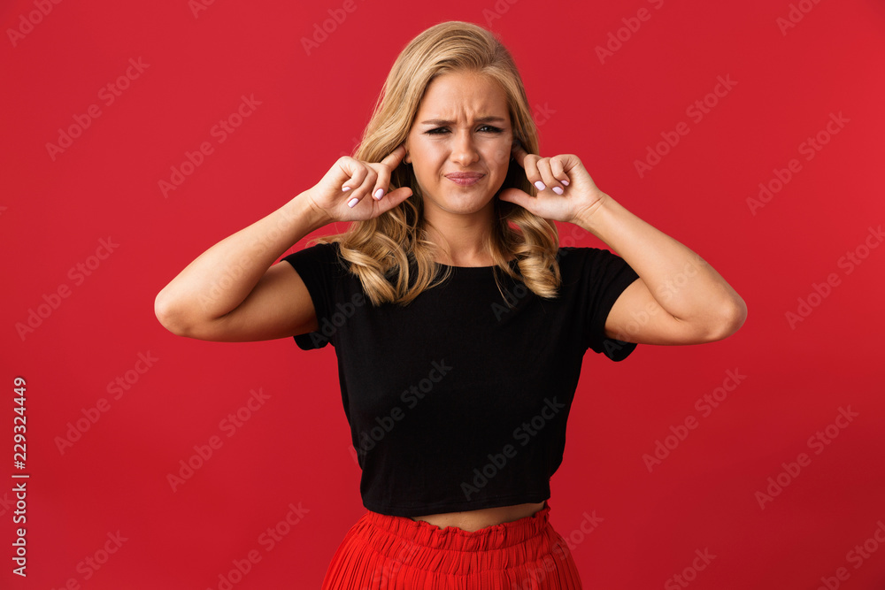 Image of displeased blonde woman 20s covering ears with hand, isolated over red background in studio Image of displeased blonde woman 20s covering ears with hand, isolated over red background in studio